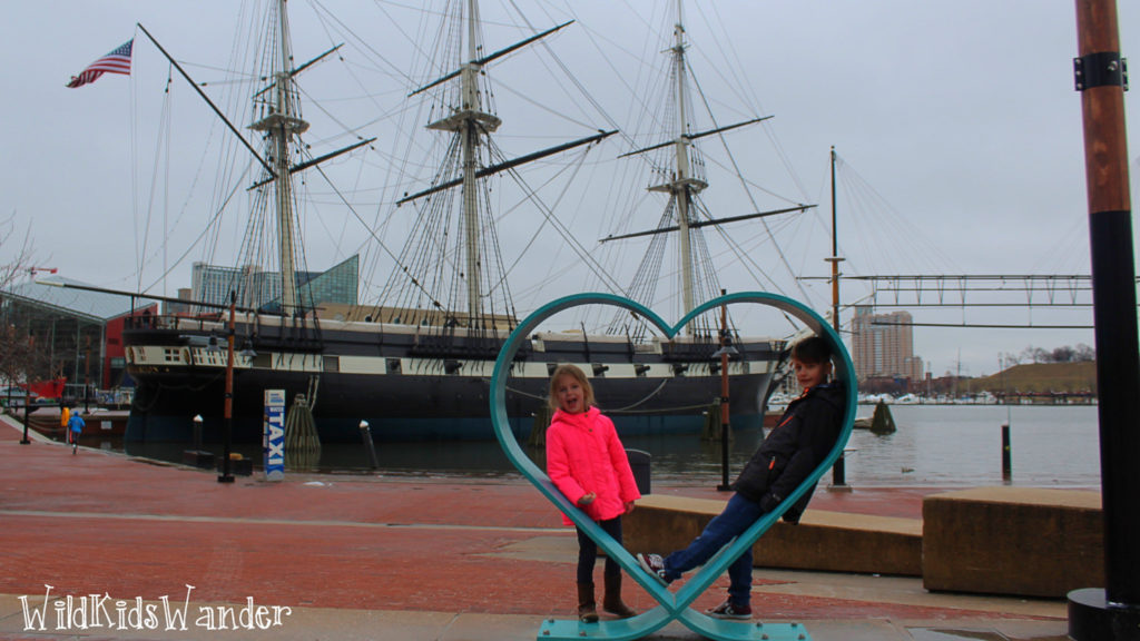 two children sit in a heart sculpture at Baltimore's Inner Harbor with a historic ship in the background