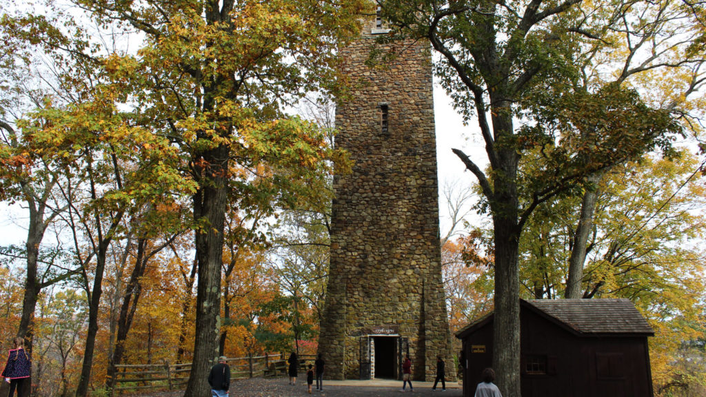 A large stone tower stands among tall trees in the fall while people walk around its base