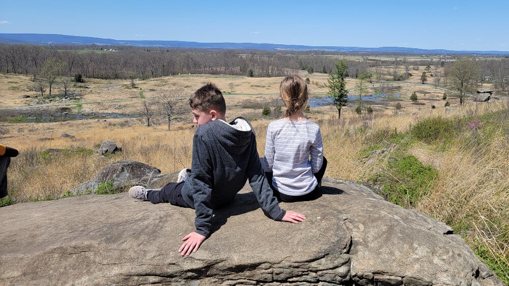 Kids will love exploring the battlefield at Gettysburg. Photo shows two kids sitting at Little Round top looking over Gettysburg battlefield.