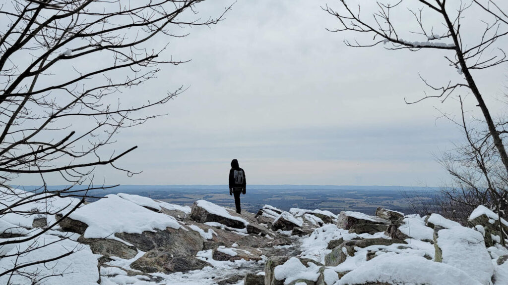 A young man stands at an overlook with snow and rocks on the ground.