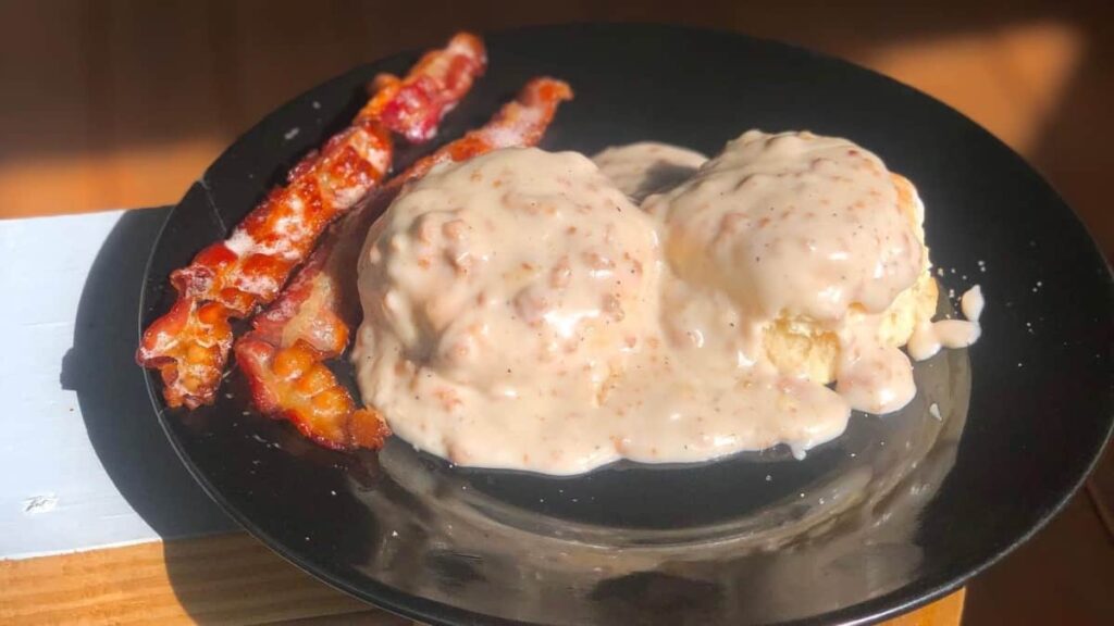 on a black plate, two biscuits are covered in a sausage cream sauce with bacon on the side
