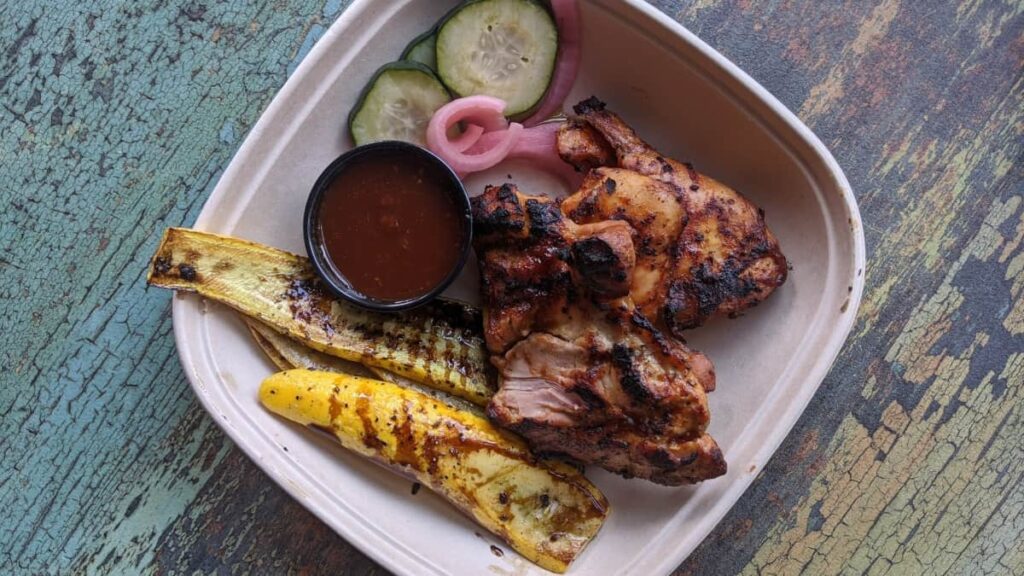 on a square paper plate, grilled yellow squash and two grilled chicken thights with barbecue sauce in a black cup on the side