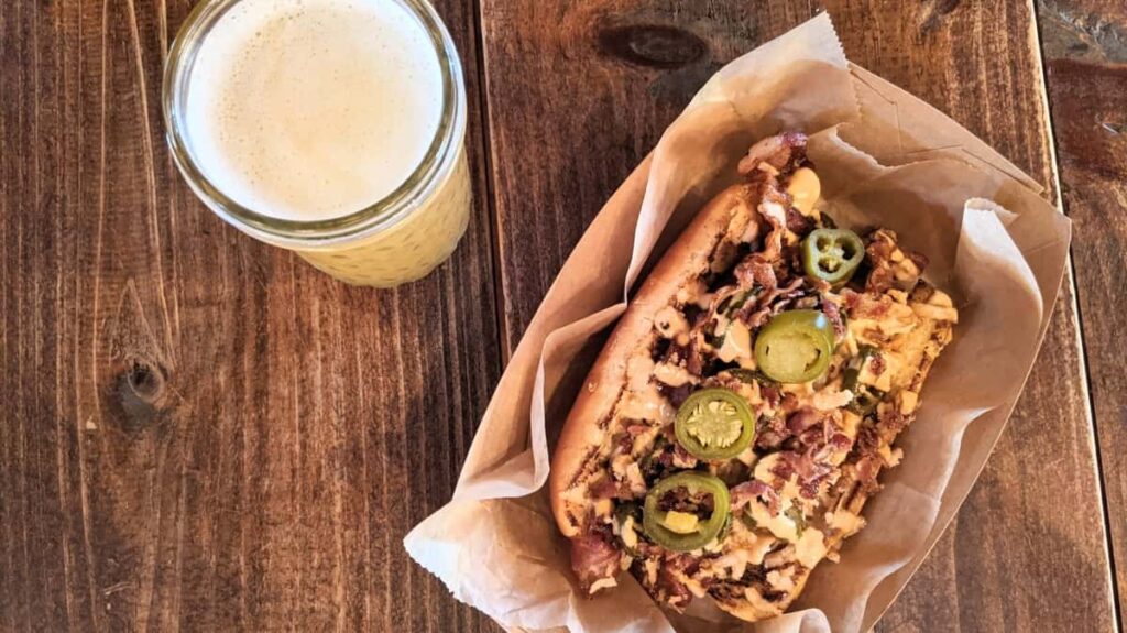 a overhead look at a beer and a hot dog covered in peppers, cheese, and bacon