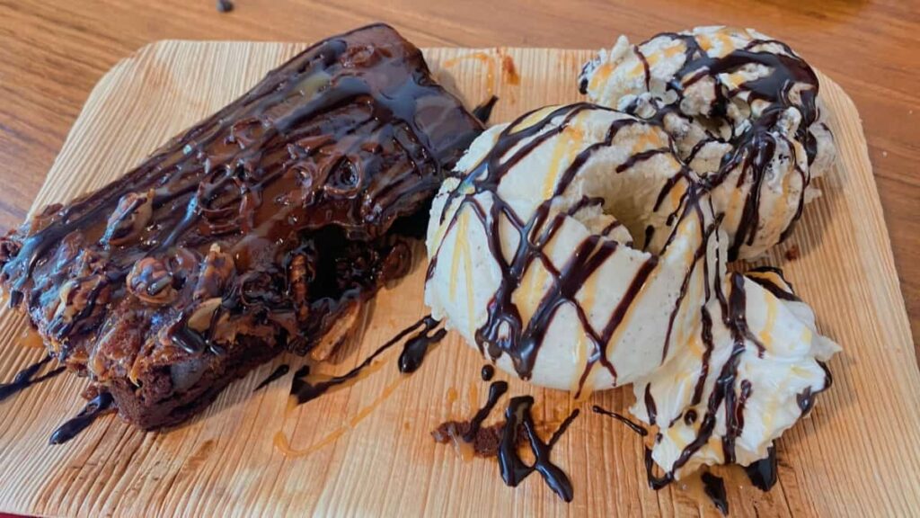 a wooden plate holds a brownie and scoop of vanilla ice cream with drizzled chocolate syrup on top