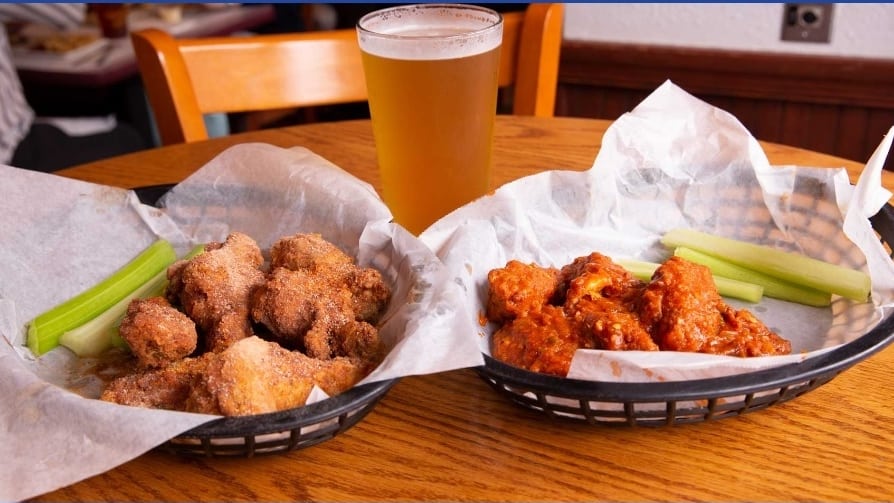 two food baskets sitting on a restaurant table with buffalo wings inside and a beer glass in the back