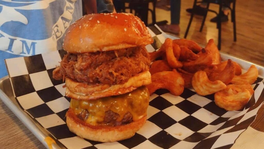 A double stacked pulled pork sandwich sits on black and white checkered paper with french fries in the back
