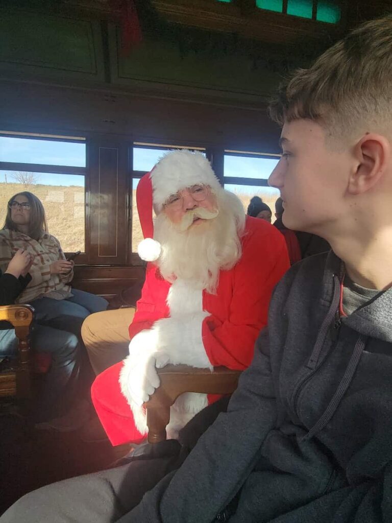 Santa claus crouches down in a train aisle to speak with a teenage boy